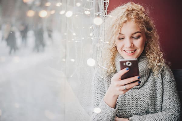 Mergina su išmaniuoju telefonu prie lango su girlianda